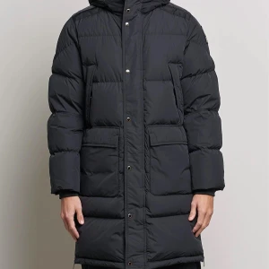 Svart lång dunjacka från Parajumpers - Säljer en svart lång dunjacka från Parajumpers med huva och knäppning framtill. Jackan har en broderad logga på ärmen och stora fickor. Perfekt för kalla dagar. Ny pris 9990kr.