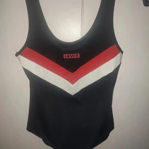 Svart baddräkt från Levi's - Snygg svart baddräkt från Levi's med ett coolt V-format mönster i rött och vitt på framsidan. Perfekt för en dag på stranden eller vid poolen. Den har en klassisk passform och breda axelband för extra komfort.