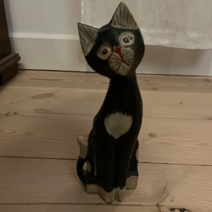 Dekorativ träkatt - Charmig handgjord träkatt med svart kropp och vita detaljer. Katten har en söt, snedställd huvudposition och ett uttrycksfullt ansikte med röda och vita detaljer. Perfekt som prydnad i hemmet för kattälskare. Höjd 28 cm. Har totalt 4 st i olika storlekar 