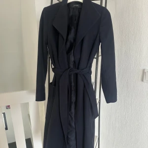 Svart kappa från Zara Basic - Elegant svart kappa från Zara Basic med knytband i midjan. Perfekt för att hålla stilen under kyligare dagar. Klassisk design med långa ärmar och en stilren look.