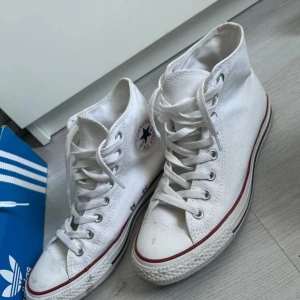 Vita Converse All Star sneakers - Säljer ett par klassiska vita Converse All Star sneakers med hög modell och snörning. Skorna har den ikoniska röda och blå loggan på sidan och en röd rand runt sulan. Perfekta för en avslappnad stil.   Storlek 41