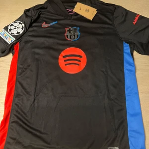 Svart FC Barcelona fotbollströja - Snygg svart FC Barcelona tröja med röda och blå detaljer. Kortärmad design med klubbmärke och sponsorlogga på framsidan. Champions League-märke på ärmen och spelarnamn och nummer 19 på baksidan.