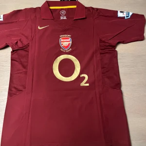 Vinröd Arsenal fotbollströja - Säljer en vinröd Arsenal fotbollströja från Nike med korta ärmar. Tröjan har klubbens emblem och sponsorlogga i guld på framsidan samt spelarnamnet och numret 'Henry 14' på baksidan. Perfekt för samlare eller fans av Arsenal.