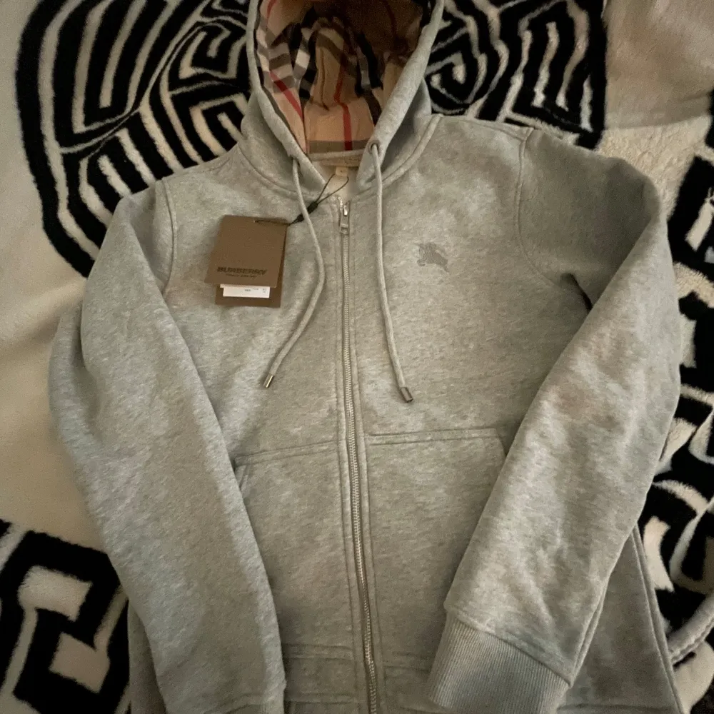 Säljer en stilren helt ny oanvänd grå hoodie från Burberry med dragkedja och broderad logga på bröstet. Insidan av huvan har det klassiska Burberry-mönstret. Perfekt för en avslappnad och trendig look. om du vill ha fler bilder så är det bara att säga till. Neuletakit & Villapaidat.