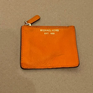 Michael Kors Coin Pouch Myntbörs - Mått: 9,3 cm x 7,2 cm. YKK dragkedjan ger både funktion och kvalitet, medan de subtila tecknen på användning, som ett litet skavt hörn och ett knappt synligt streck på framsidan, inte stör den eleganta designen. En perfekt liten väska för att förvara dina små värdesaker med både stil och finess.