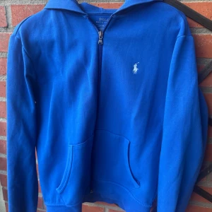 Blå hoodie från Polo Ralph Lauren - Säljer en snygg blå hoodie från Polo Ralph Lauren med dragkedja och det klassiska logotypen på bröstet. Perfekt för en avslappnad stil. Den har långa ärmar och en praktisk huva. Nypris ca 2200