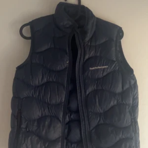 Svart dunväst från Peak Performance - Snygg svart dunväst från Peak Performance med dragkedja framtill. Perfekt för kyligare dagar och ger en sportig look. Västen har en quiltad design som ger extra värme och stil. (Defekt på fickan)