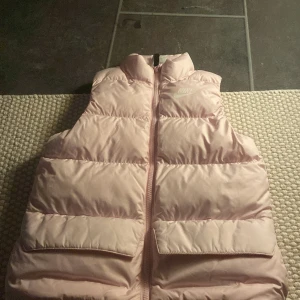 Rosa dunväst från Nike - Säljer en snygg rosa dunväst från Nike. Västen har en dragkedja framtill och två praktiska fickor. Perfekt för kyliga dagar när du vill hålla värmen utan att kompromissa med stilen.