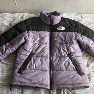 Lila och svart pufferjacka från The North Face - Säljer en lila och svart pufferjacka från The North Face. Jackan har en dragkedja framtill och två fickor med dragkedjor. Perfekt för kyligare dagar med sin vadderade design. Logotypen är broderad på bröstet och ryggen.