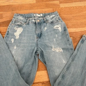 Blå slitna jeans - Säljer ett par blå jeans med slitningar och hål för en trendig look. De har en hög midja och en straight passform. Perfekta för en avslappnad stil.
