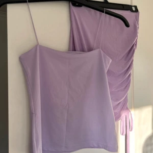 Ljuslila kjol med tillhörande linne - Snygg ljuslila kjol med drapering på sidan som ger en fin detalj. Kjolen har en mjuk och följsam passform, perfekt för en stilren look. Den är gjord i ett lätt material som faller fint. Tillhörande linne med.