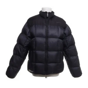 Svart pufferjacka från Nike - Säljer en svart pufferjacka från Nike i storlek XL. Jackan har en dragkedja framtill och är tillverkad av nylon och polyester. Perfekt för kalla dagar med sin vaddering av dun och fjädrar. Den har en stilren design med Nike-loggan på bröstet.
