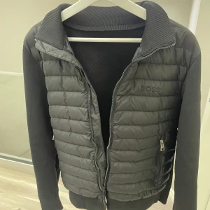 Cardigan - Snygg svart cardigan jacka från Boss med quiltad framsida och ribbade ärmar. Jackan har en dragkedja framtill och en diskret logga på bröstet samt en etikett på ärmen. Perfekt för en stilren look.