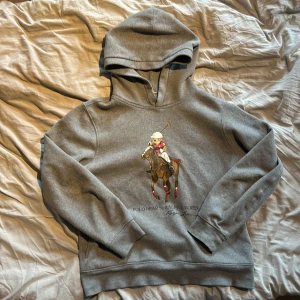 Grå hoodie från Ralph Lauren - Säljer en grå hoodie från Ralph Lauren med ett stort tryck av en polospelande björn på framsidan. Tröjan har långa ärmar och en klassisk huva. Perfekt för en avslappnad stil.