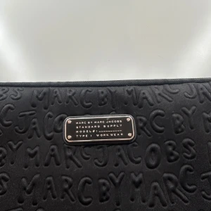 Svart datorväska från Marc by Marc Jacobs - Snygg svart datorväska från Marc by Marc Jacobs med präglad logotypdesign. Väskan har en dragkedja för säker förvaring av din laptop. Perfekt för att bära med sig datorn med stil. Stl 13. Lite färg har lossnat på dragkedjan