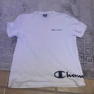 Vit t-shirt från Champion - Säljer en vit t-shirt från Champion med deras logga tryckt både på bröstet och nedtill. T-shirten har en klassisk design med rund hals och korta ärmar. Perfekt för en avslappnad stil.