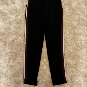 Svarta byxor med ränder från Zara - Eleganta svarta byxor från Zara med en sportig touch. Byxorna har en stilren design med en dekorativ rand längs sidan i färgerna vinröd och vit. Perfekt för en avslappnad men ändå sofistikerad look.  Strl: 38 Skick: bra skick Ord. Pris: 399 Säljes då de blivit små på mig  Din stil: spara inlägget så du inte missar denna kappa Inte din stil: Kolla min profil. Du kommer hitta något för alla kön och åldrar.