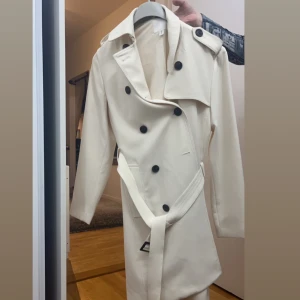 Beige trenchcoat från H&M - Snygg beige trenchcoat från H&M med klassisk design. Jackan har långa ärmar och knappar framtill samt ett bälte i midjan för en stilren look. Perfekt för övergångsperioder.
