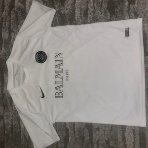 Vit fotbollströja från Nike x Balmain - Snygg vit fotbollströja från Nike i samarbete med Balmain. Tröjan har korta ärmar och ett stilrent tryck med Balmain Paris-logga på bröstet. Perfekt för dig som vill ha en exklusiv look på planen eller till vardags.