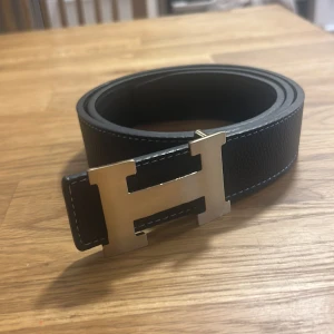 H bälte - Snyggt svart bälte med ett stort H-format spänne i silver. Tillverkat i skinn med en stilren design och diskreta sömmar längs kanterna. Perfekt för att ge en elegant touch till din outfit.  Storleken är 44/110