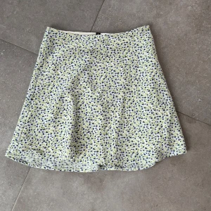 Blommig kortkjol - Söt blommig kortkjol i ljusa färger med små blå och gula blommor. Perfekt för varmare dagar och ger en fräsch och somrig look. Kjolen har en enkel och stilren design som gör den lätt att matcha med olika toppar.