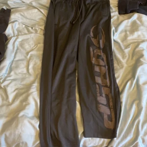 Gråa gymbyxor från Gasp - Säljer ett par svarta mjukisbyxor från Gasp med elastisk midja och snörning. Byxorna har en stor logga längs benet och är perfekta för en avslappnad stil. De är tillverkade i ett mjukt material som ger en bekväm passform.