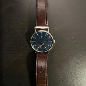  klocka från Carl Edmond - Stilren klocka med en djupblå urtavla och brunt skinnarmband. Klockan har en silverfärgad boett och tydliga markeringar. Perfekt för den som söker en klassisk och tidlös design. Nypris 2000kr