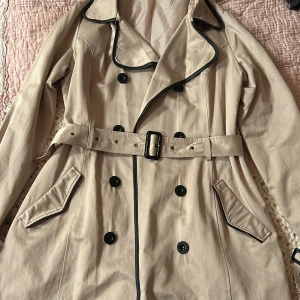 Beige trenchcoat från Oxmo - Snygg stilren beige trenchcoat från Oxmo med svarta detaljer och knappar. Jackan har ett bälte i midjan och klassisk krage. Perfekt för höst och vår. En slayig version av Sherlock Holms helt enkelt✨Become the mystery sis 🕵️