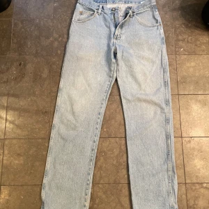 Ljusblå jeans från Wrangler - Säljer ett par klassiska ljusblå jeans från Wrangler. De har en straight passform och är tillverkade i slitstarkt denim. Perfekta för en avslappnad vardagslook. Jeansen har fem fickor och en knappgylf.