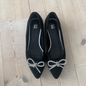 Svarta ballerinaskor med glittrig rosett från XIT - Säljer ett par eleganta svarta ballerinaskor från XIT men köpa på din sko. De har en spetsig tå och är dekorerade med en glittrig rosett framtill. Perfekta för både fest och vardag! Skorna är i nyskick och redo att användas.