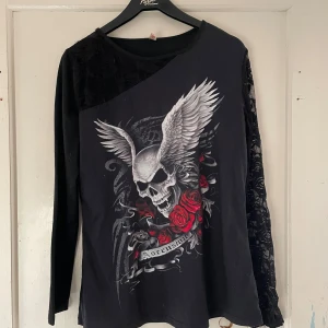 Svart långärmad tröja med dödskallemotiv - Säljer en cool svart långärmad tröja med ett stort tryck av en dödskalle med vingar och röda rosor. Ena ärmen har ett snyggt spetsmönster som ger en unik touch. Perfekt för höst och vinter när du vill ha något både bekvämt och stiligt. Passar bra till både vardag och fest!