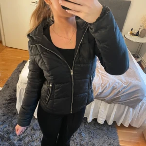 Svart pufferjacka med dragkedja - Säljer en snygg svart pufferjacka som är perfekt för små kyliga dagar. Jackan har en dragkedja framtill och två fickor med dragkedjor. Den är långärmad och har en huva som skyddar mot vind och regn. Passar perfekt för höst och vinter!