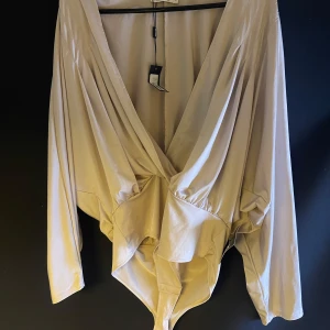 Beige omlottbody - Säljer en superfin beige omlottbody med långa ärmar. Den har en djup v-ringning och är perfekt för både vardag och fest. Materialet känns mjukt och skönt mot huden. Passar perfekt till höst- och våroutfits!