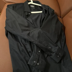 Svart skjorta från Dressmann - Snygg svart skjorta från Dressmann i slim fit. Tillverkad i premium bomull med stretch för extra komfort. Skjortan har långa ärmar och knappar framtill. Perfekt för en stilren look.