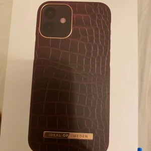 Brun mobilskal från Ideal of Sweden - Snyggt mobilskal i brun krokodilimitation från Ideal of Sweden. Skalet har en elegant design med en guldig detalj vid kameran och logotypen. Perfekt för att ge din telefon ett stilrent och lyxigt utseende.