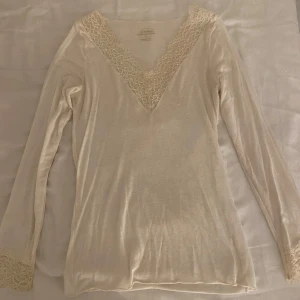 Beige/Vit långärmad topp från Intimissimi - Säljer en elegant beige/Vit långärmad topp från Intimissimi. Toppen har en vacker spetsdetalj vid v-ringningen och ärmsluten. Den är sällan använd och är i bra skick