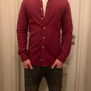 Vinröd skjorta Ralph lauren - Snygg vinröd skjorta från Ralph Lauren med klassisk krage och knappar framtill. Skjortan har långa ärmar och en liten broderad logga på bröstet. Perfekt för en stilren look.