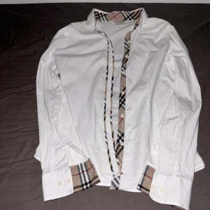 Vit skjorta från Burberry - Säljer en stilren vit skjorta från Burberry med klassiskt rutigt mönster på insidan av kragen och knappslån. Skjortan har långa ärmar och knappar vid ärmsluten. Perfekt för en elegant look.