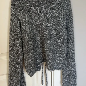 Grå stickad tröja från NA-KD - Säljer en mysig grå stickad tröja från NA-KD med en unik öppen rygg och knytning. Perfekt för kyliga dagar med sin långa ärmar och mjuka material. Tröjan har en melerad look som ger en trendig touch.