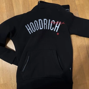 Svart hoodie från Hoodrich - Säljer en svart hoodie från Hoodrich med stor text på ryggen och detaljer på huvan. Perfekt för en casual look med känguruficka framtill. Huvtröjan har en stilren design med broderade detaljer. Den är i väldig bra skick 