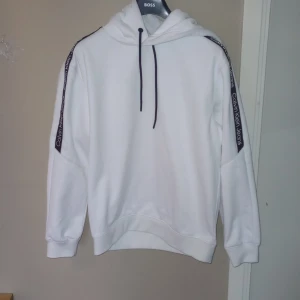 Vit hoodie från Calvin Klein Jeans - Säljer en stilren vit hoodie från Calvin Klein Jeans med svarta snören och logotyp längs ärmarna. Hoodien har en klassisk passform och är tillverkad i mjukt material för extra komfort. Hoodien är i storlek M. 