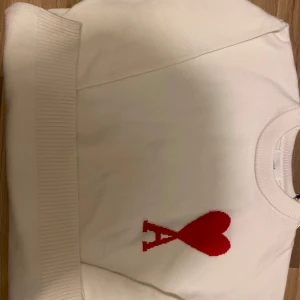 Vit tröja med rött hjärtmotiv från Ami - Säljer en stilren vit tröja från Ami med ett stort rött hjärtmotiv och bokstaven 'A' på framsidan. Tröjan har en rund halsringning och ribbade muddar vid ärmslut och nederkant. Perfekt för en trendig look! Oanvänd säljer pga att den inte passar mig (Size m) med orginal förpackning och fungerande Scan 