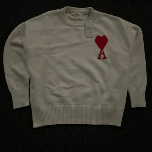 Vit tröja från Ami Alexandre Mattiussi - Säljer en stilren vit tröja från Ami Alexandre Mattiussi med ett rött hjärta och bokstaven 'A' broderat på bröstet. Den är helt oanvänd för stor storlek bara för mig.