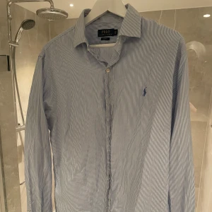 Randig skjorta från Ralph Lauren - Snygg och stilren blå- och vitrandig skjorta från Ralph Lauren. Skjortan har en klassisk krage och knappar framtill. Perfekt för en elegant look med sin slim fit passform.