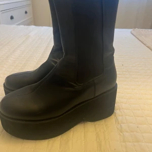 Svarta platåstövlar - Snygga svarta platåstövlar i skinn med en hög sula. De har en stilren design med elastiska paneler på sidorna för enkel på- och avtagning. Perfekta för att ge lite extra höjd och stil till din outfit.
