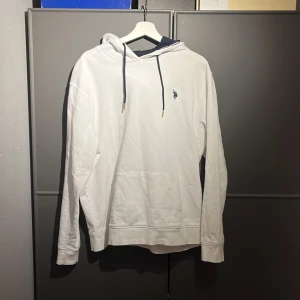 Vit hoodie från U.S. Polo Assn. - Säljer en stilren vit hoodie från U.S. Polo Assn. med en liten broderad logga på bröstet. Hoodien har långa ärmar och en klassisk känguruficka framtill. Tillverkad i mjuk bomull för extra komfort. Den här för det mesta hängt i min garderob inte använd på ett bra tag men inga större fel på den