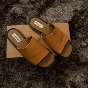 Bruna sandaler från Steve Madden - Snygga bruna sandaler från Steve Madden med en bred rem över foten. Sandalerna har en stilren design med en platt sula och öppen tå, perfekt för varma dagar.