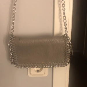 Grå axelväska från Tiamo - Snygg grå axelväska från Tiamo med kedjedetaljer runt kanterna och som axelrem. Perfekt för att ge en stilren touch till din outfit. Väskan har en praktisk storlek för vardagliga behov.