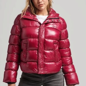 Röd pufferjacka - Säljer en snygg röd pufferjacka från superdry med dragkedja och knappar. Jackan har en glansig finish och långa ärmar, perfekt för kyliga dagar. Den är både stilren och praktisk med fickor framtill. 