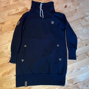 Svart knälång collegetröja från Tindra - Säljer en svart knälång collegetröja från Tindra med hög krage och snörning. Tröjan har långa ärmar och praktiska fickor framtill. Perfekt för kyliga dagar.